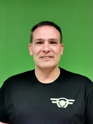 Porträt eines lächelnden Mannes vor grünem Hintergrund, trägt ein schwarzes T-Shirt mit einem stilisierten Logo auf der Brust.