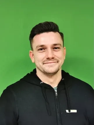 Junger Mann mit kurzem dunklem Haar und schwarzem Kapuzenpullover lächelt vor grünem Hintergrund.