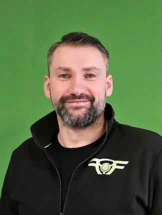 Mann mit dunklem T-Shirt und Jacke vor grünem Hintergrund, der freundlich lächelt.
