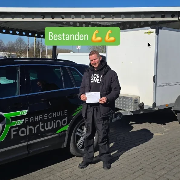 Mann steht neben Fahrschulauto und Anhänger, hält Zertifikat hoch, Text 'Bestanden' mit Muskel-Emojis über ihm.
