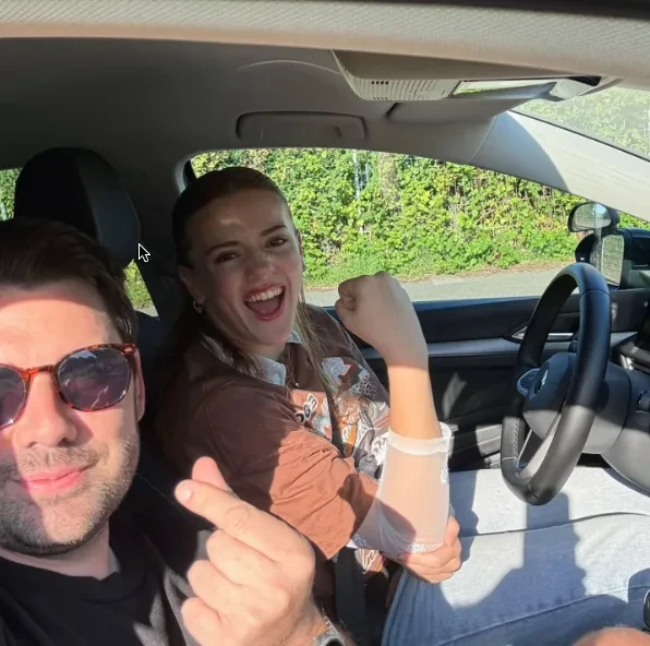 Zwei lächelnde Personen im Auto, die Kamera nach vorne zeigend, eine Person trägt eine braune Jacke.