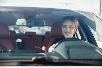 Junge Frau am Steuer eines Autos, die lächelt und durch die Windschutzscheibe sichtbar ist.
